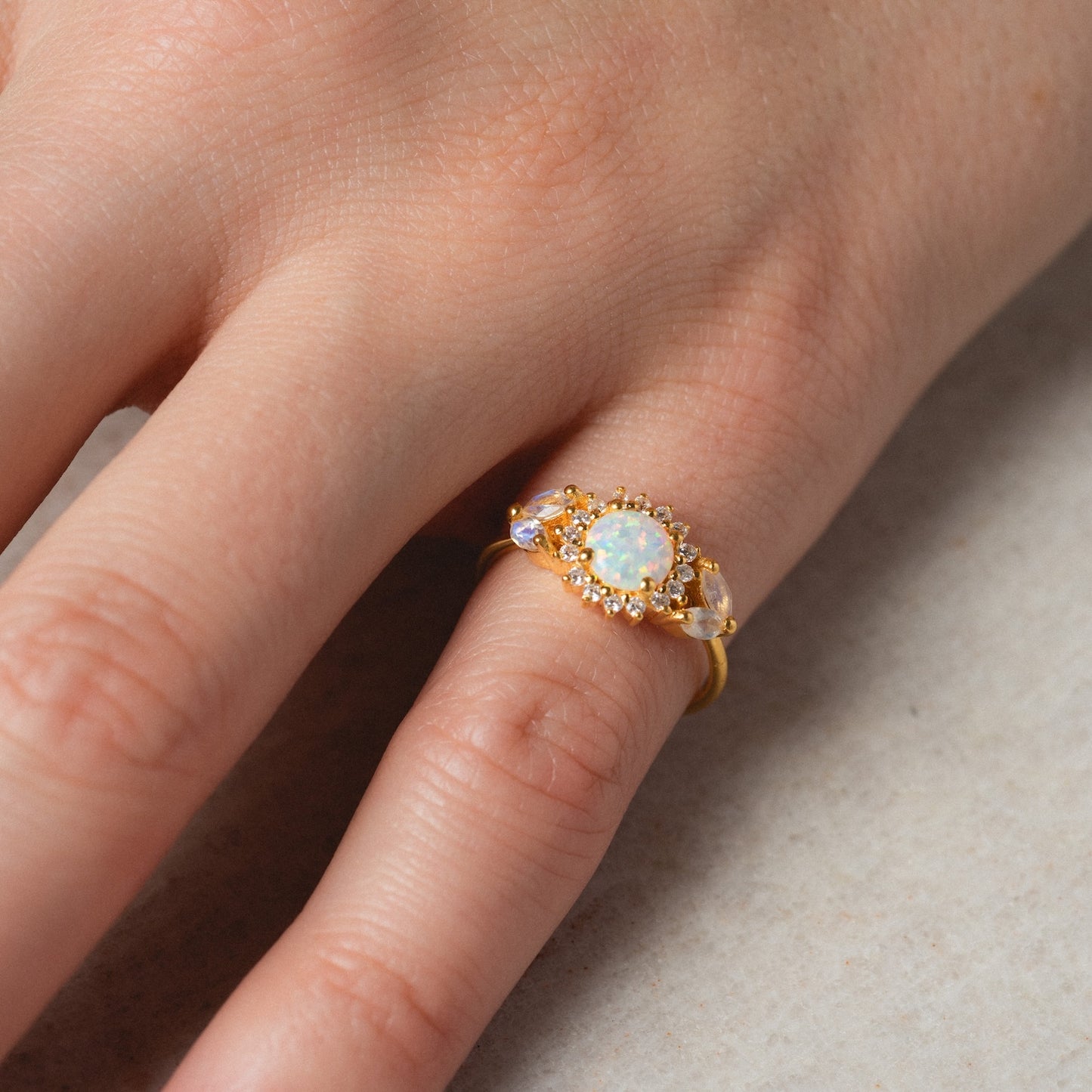 A close-up of a hand wearing the Opal and Diamond Sunflower Queen Ring, featuring a central opal stone encircled by sparkling diamonds on the ring finger against a light, textured background.