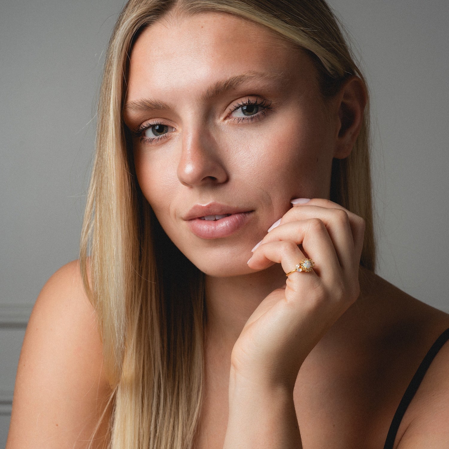 A woman with long blonde hair poses against a neutral background, resting her chin on her hand to display the Opal and Diamond Sunflower Queen Ring. She has light skin, minimal makeup, and wears a black strap.