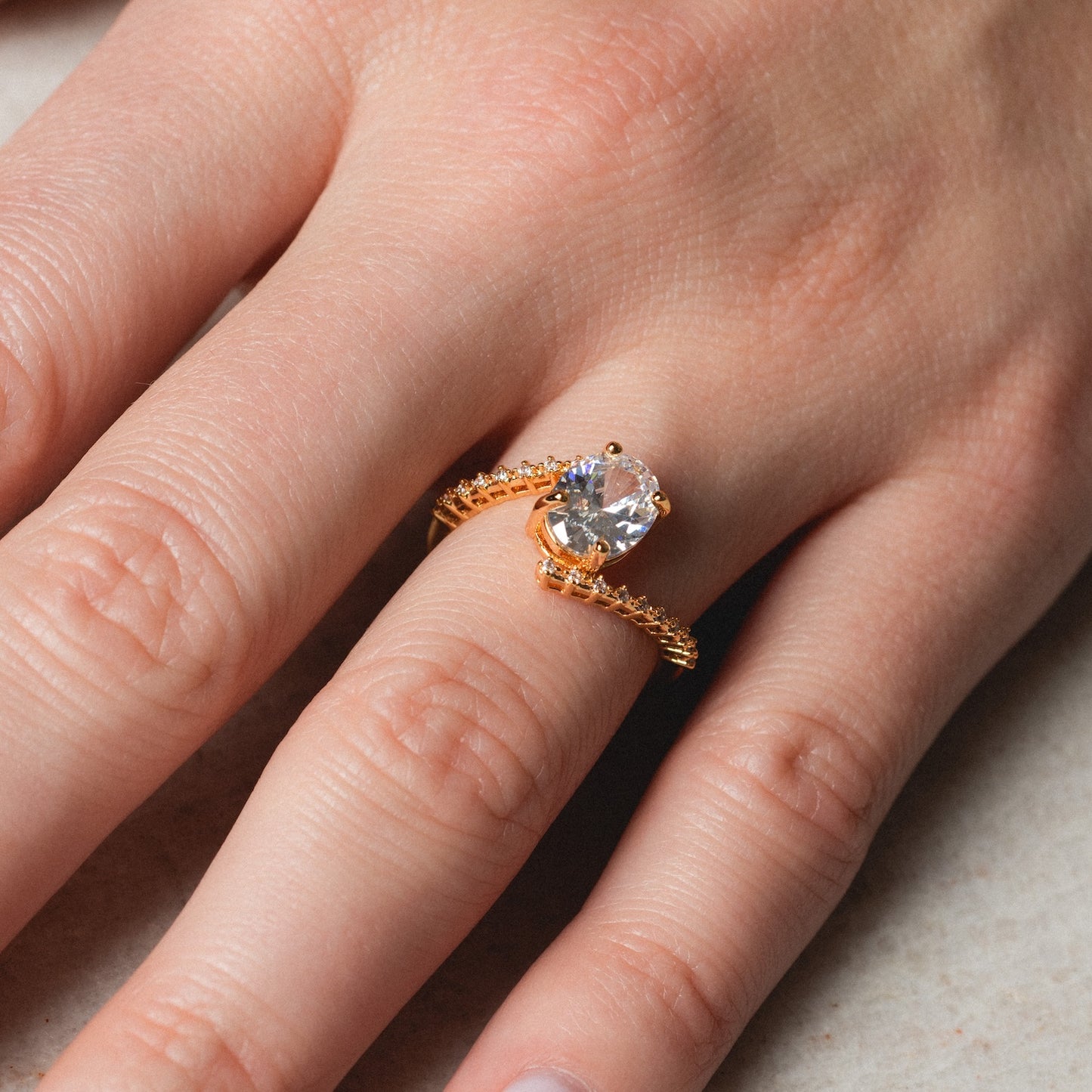 A close-up of a hand wearing the Tilted CZ Ring, featuring a large round clear cubic zirconia in a prong setting with a decorative gold band, resting on the person's ring finger.