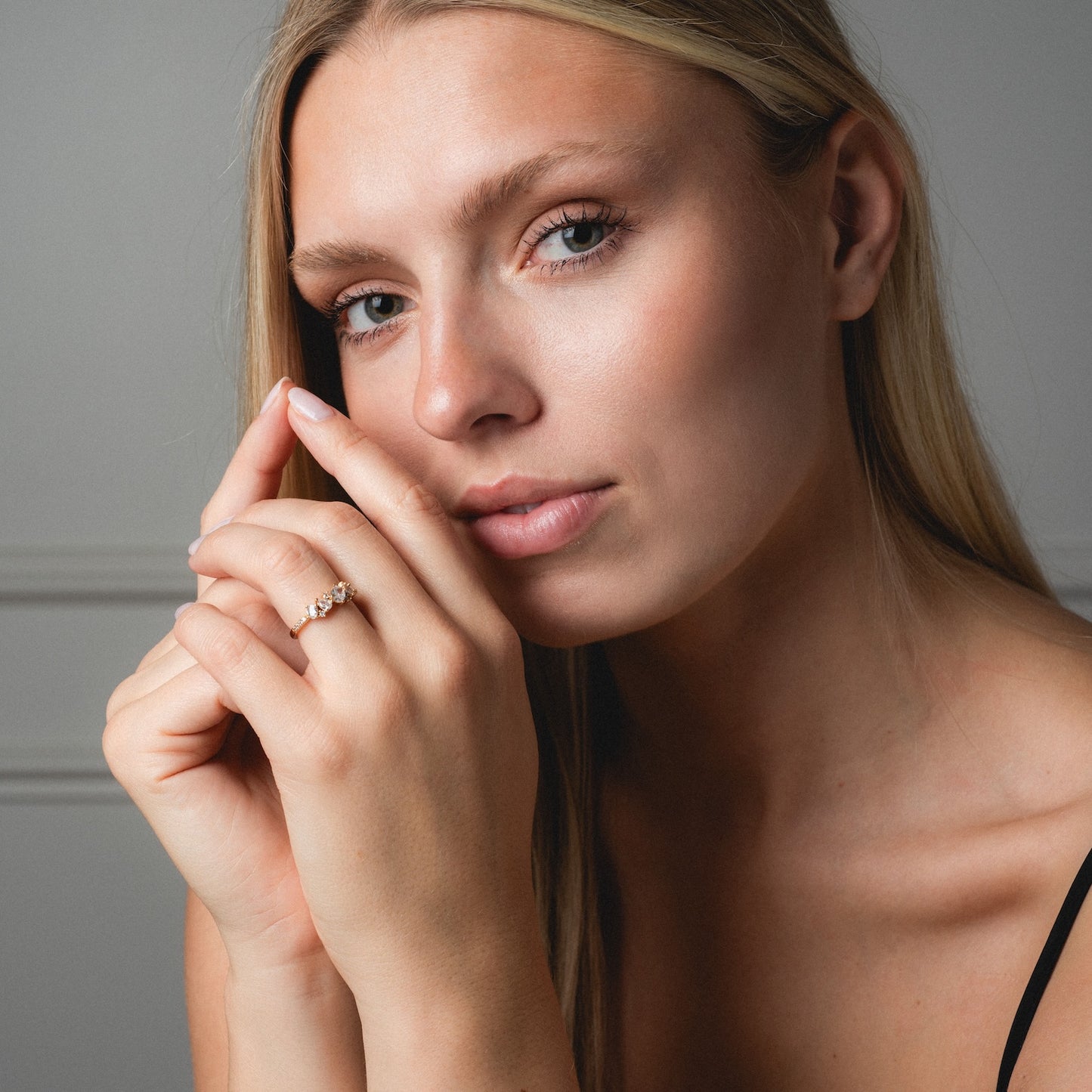 A woman with long blonde hair, wearing a black top, rests her chin on her hand and displays the Statement Crystal Cluster Ring on her finger. She has light skin and is posed against a neutral background.