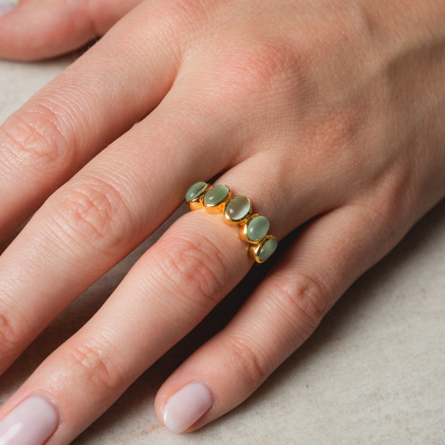 A hand with neatly manicured nails wears the Sky Blue Cats Eye Flow Ring, a gold ring featuring five oval sky blue stones arranged in a row on the middle finger, set against a light, textured background.