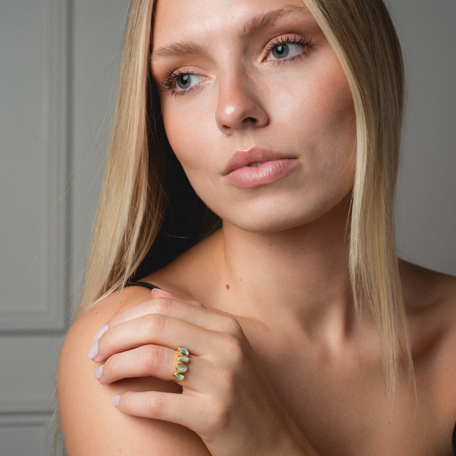 A woman with straight blonde hair and natural makeup gazes thoughtfully to the side, wearing the Sky Blue Cats Eye Flow Ring on her hand against a neutral indoor background.