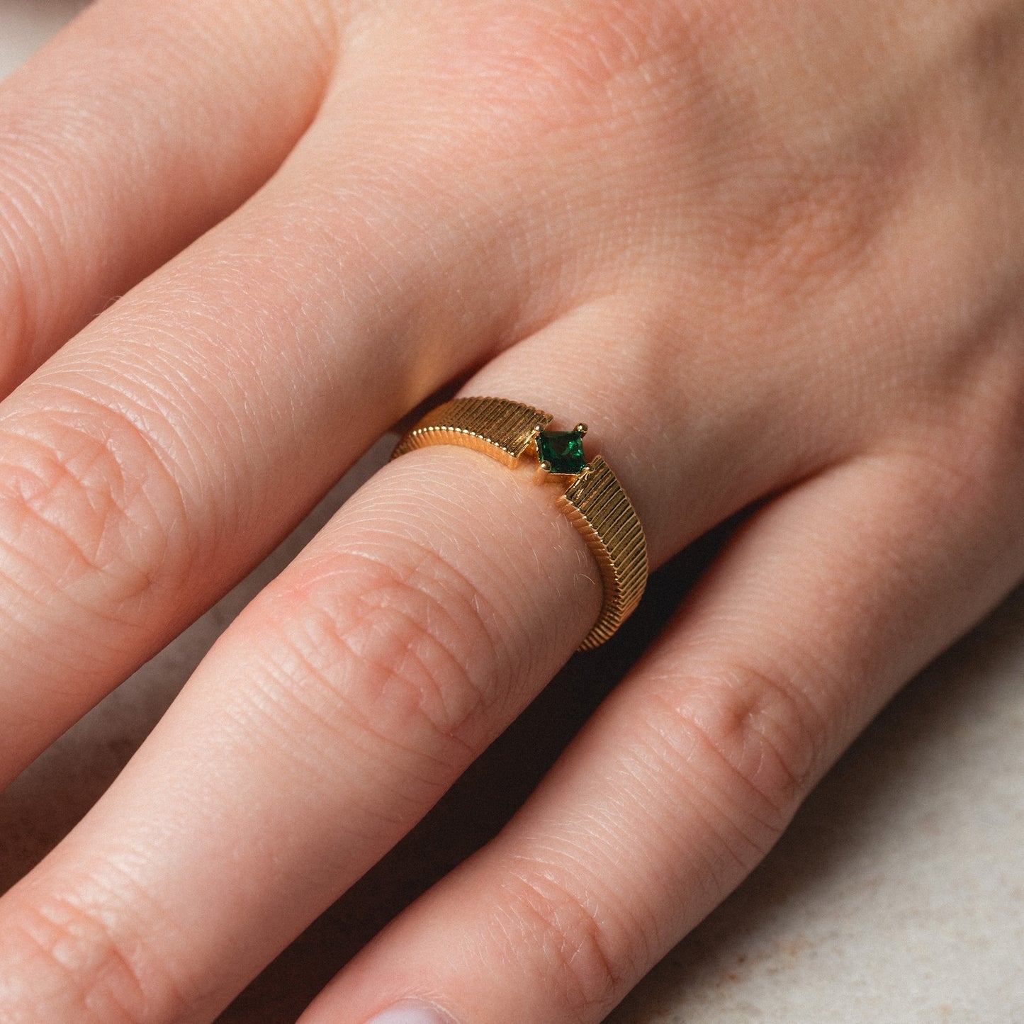 A close-up of a hand wearing the Serena Ring, a gold band featuring a small green gemstone, displayed on the ring finger against a neutral background.
