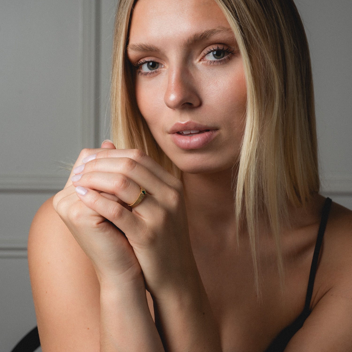 A woman with straight blonde hair and natural makeup looks at the camera, hands together by her face. She wears a thin black strap top and the Serena Ring against a neutral background.