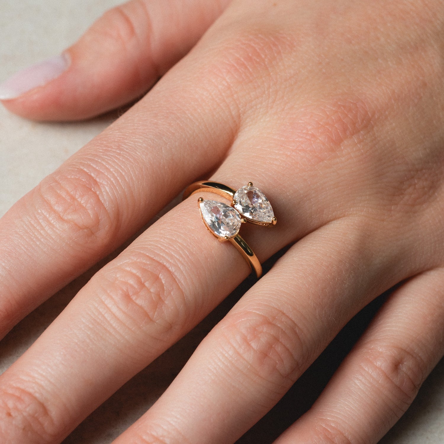 A close-up of a hand wearing the Crystal Teardrop Wrap Ring, featuring two pear-shaped clear gemstones set side by side in gold, shown against a light background.