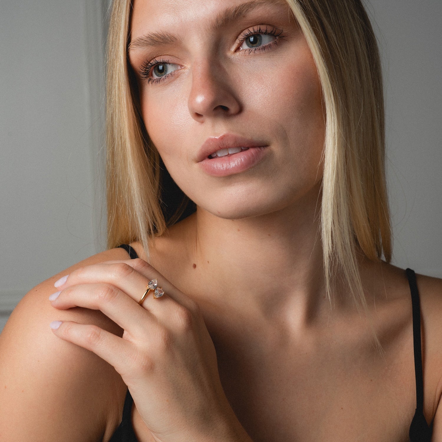 A woman with straight blonde hair and natural makeup gazes thoughtfully to the side, wearing a black top and showcasing the Crystal Teardrop Wrap Ring with a clear gemstone on her hand near her face.