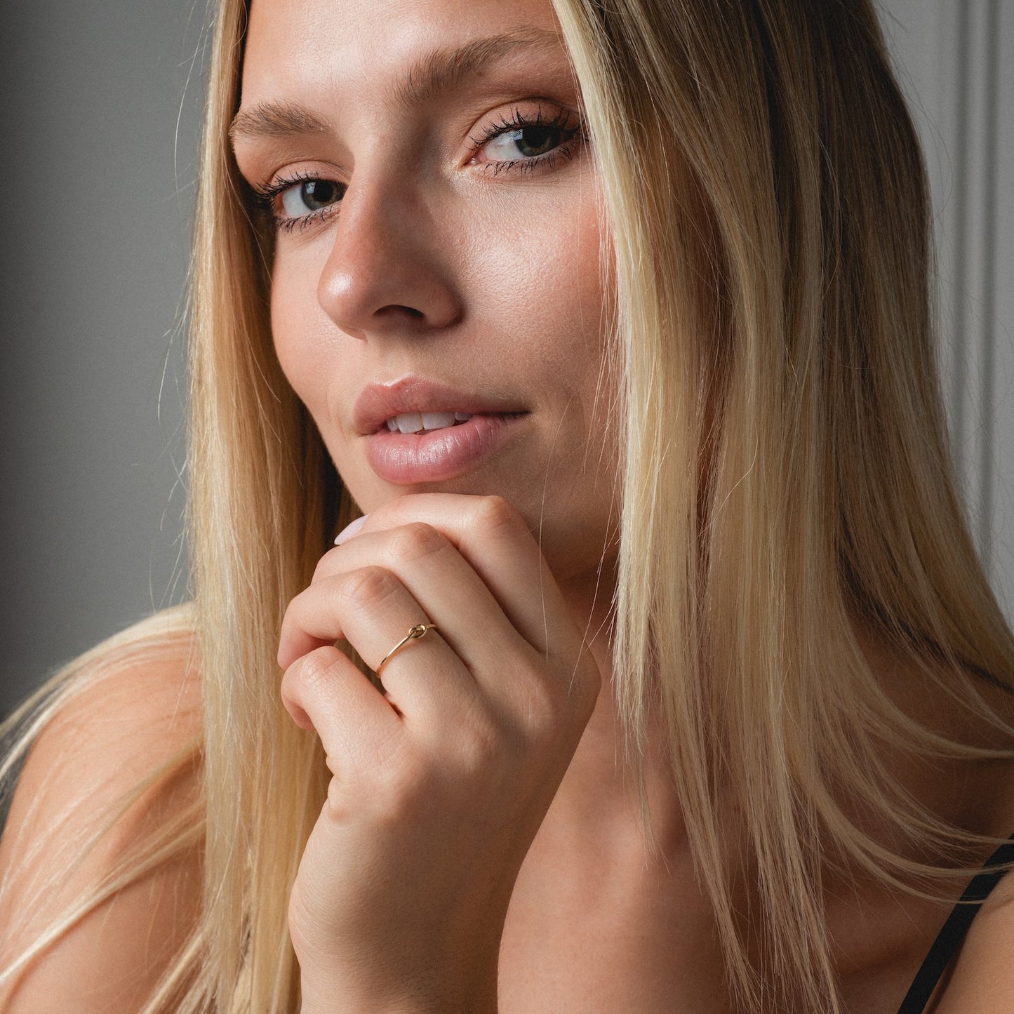A woman with long blonde hair gazes at the camera, resting her hand near her lips and wearing a Solid Gold Simple Knot Ring. The lighting highlights her natural makeup and smooth skin.