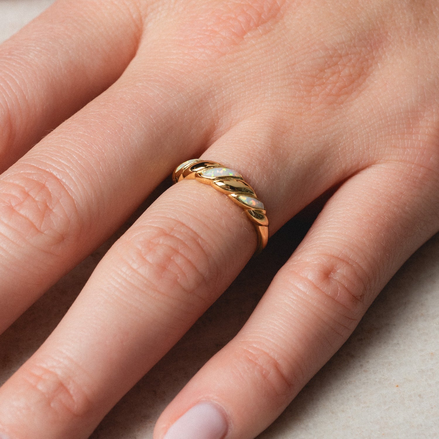 A close-up of a hand with neatly manicured nails wearing the Classic Opal Inlay Ring, a 14k gold plated vintage-inspired band, on the middle finger resting on a light surface.