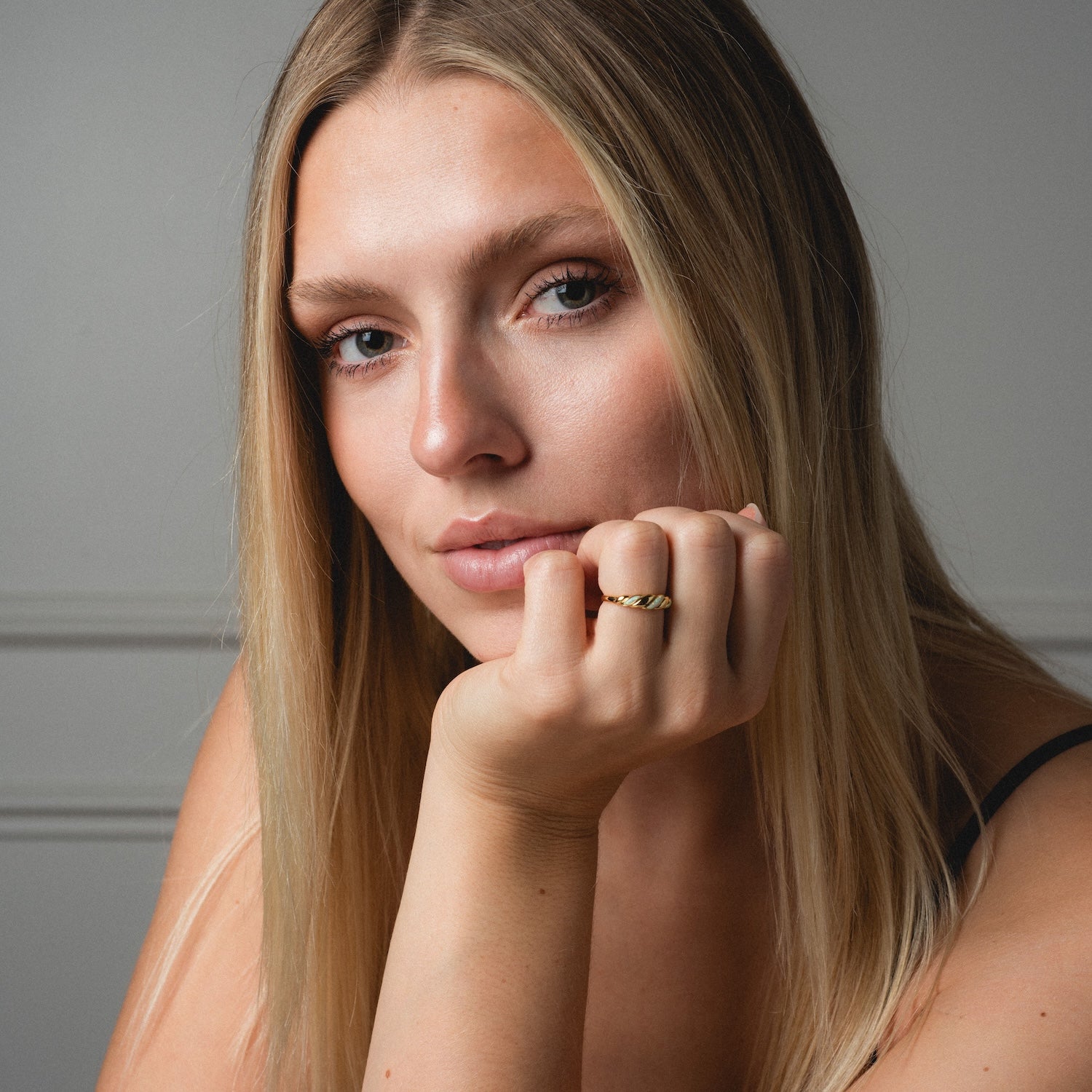 A woman with long blonde hair rests her chin on her hand, gazing at the camera. She wears the Classic Opal Inlay Ring and a black strap top, set against a soft, neutral background.