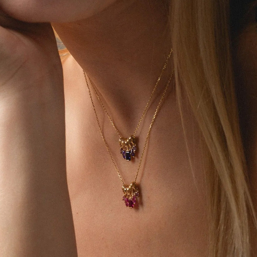 A woman with long blonde hair wears two layered Shades of You Ombre Birthstone Charm Necklaces, featuring colorful pendants, while resting her chin on her hand in a black top. The image highlights her neck and jewelry.