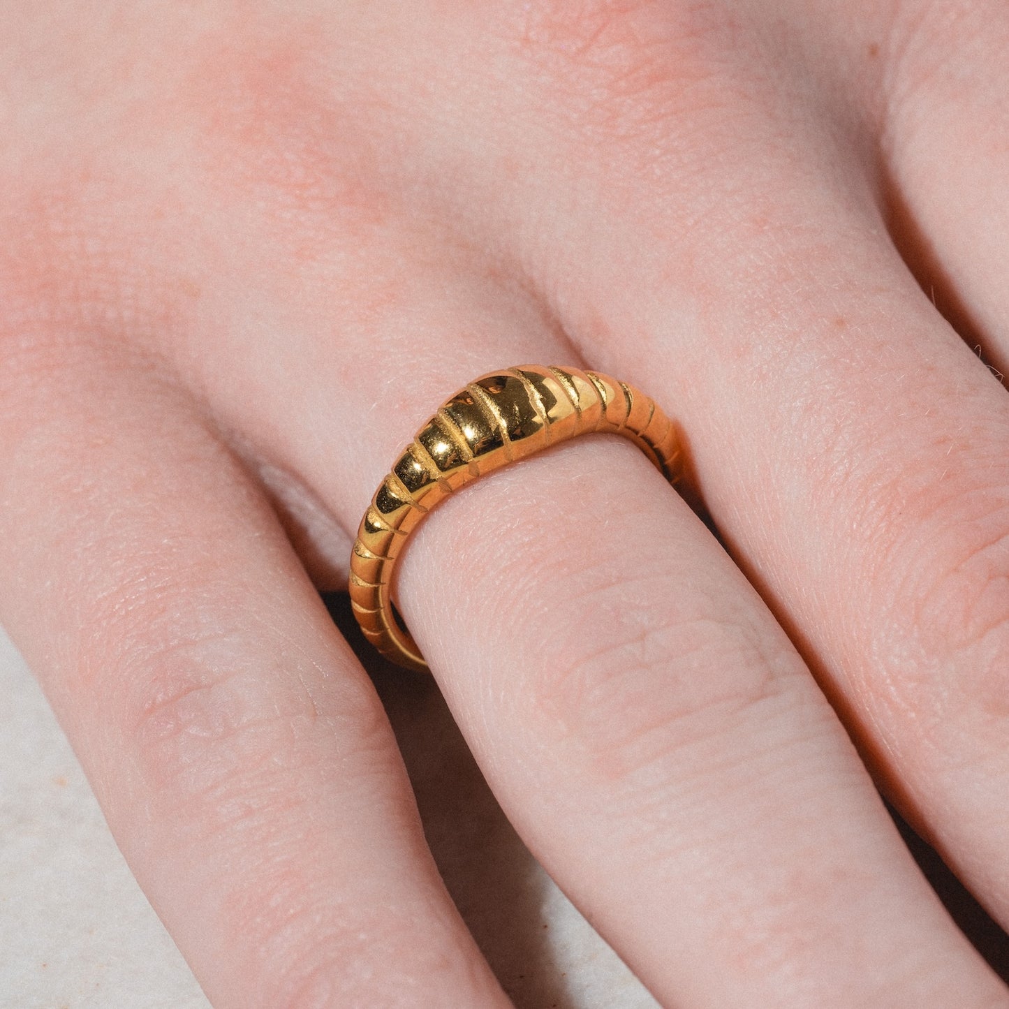 A close-up of a hand wearing the Crew Ring—a chunky, gold ring with a ridged, wave-like design and shiny finish—on the middle finger. The hand rests on a light-colored surface.