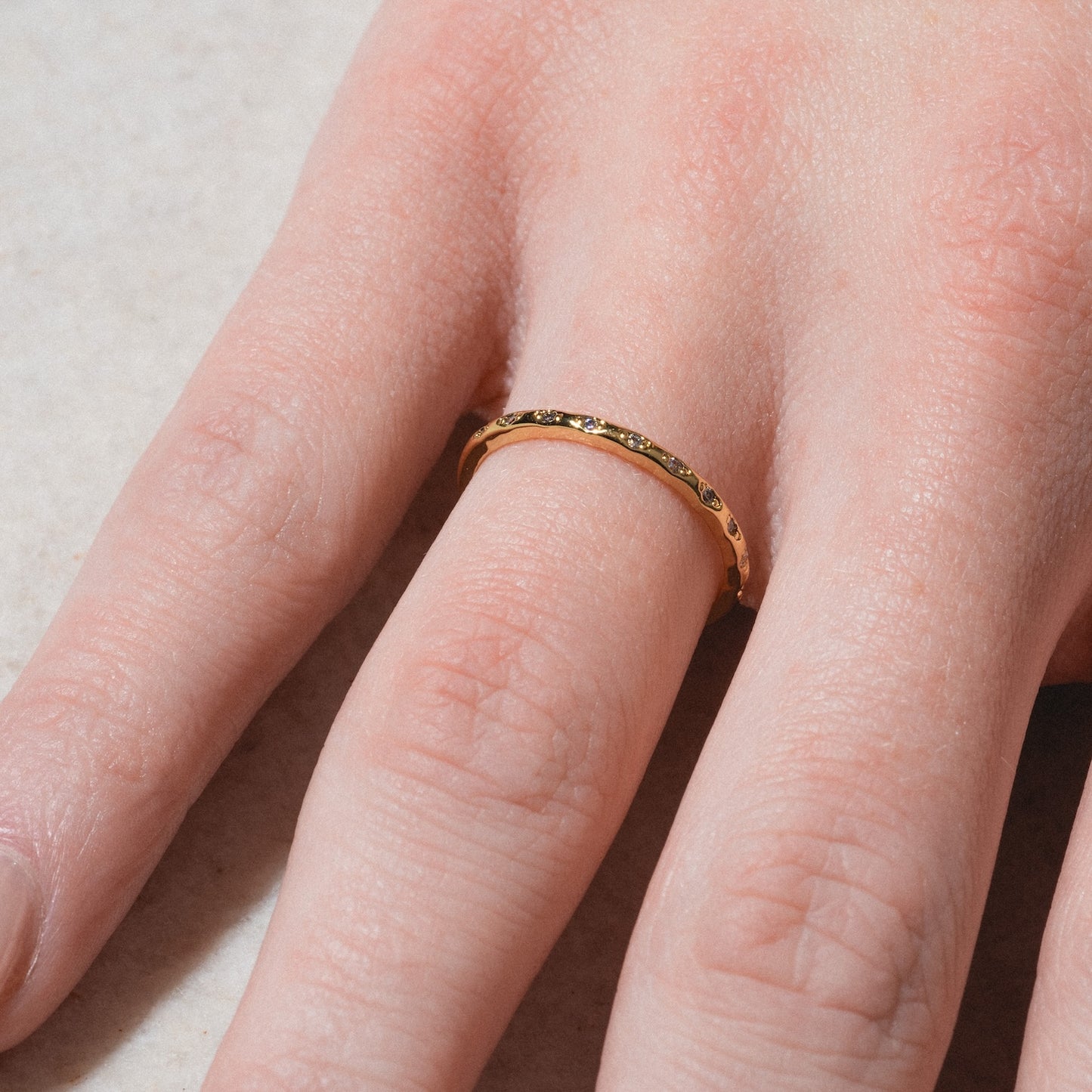 A close-up of a light-skinned hand wearing the Regency Stacking Ring, a thin textured gold-plated band on the ring finger, shown against a light, slightly speckled surface.