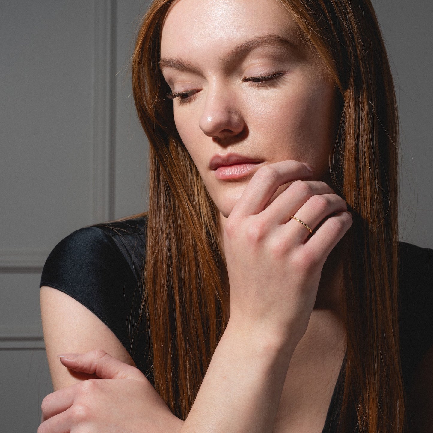 A woman with long red hair and fair skin poses with her eyes closed, gently touching her chin while wearing the Regency Stacking Ring on her finger. She is dressed in a black top against a neutral background.