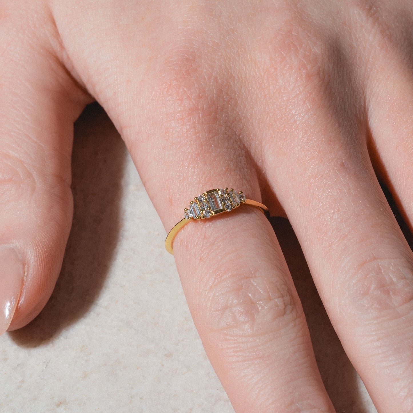 The Soiree Vintage Inspired Ring showcases a rectangular central white CZ flanked by two side stones on a 14k gold plated band, gleaming elegantly on the ring finger against a light surface.