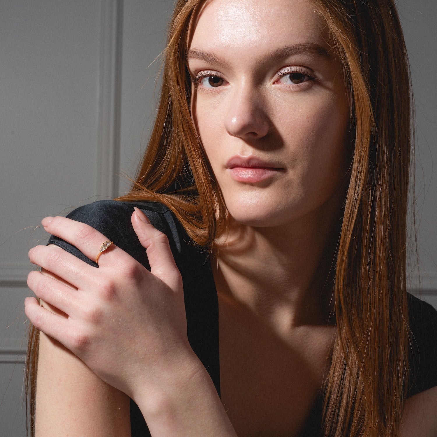 A woman with long, straight auburn hair in a black top looks at the camera with a neutral expression, her hand on her shoulder showcasing the Soiree Vintage Inspired Ring adorned with white CZ.