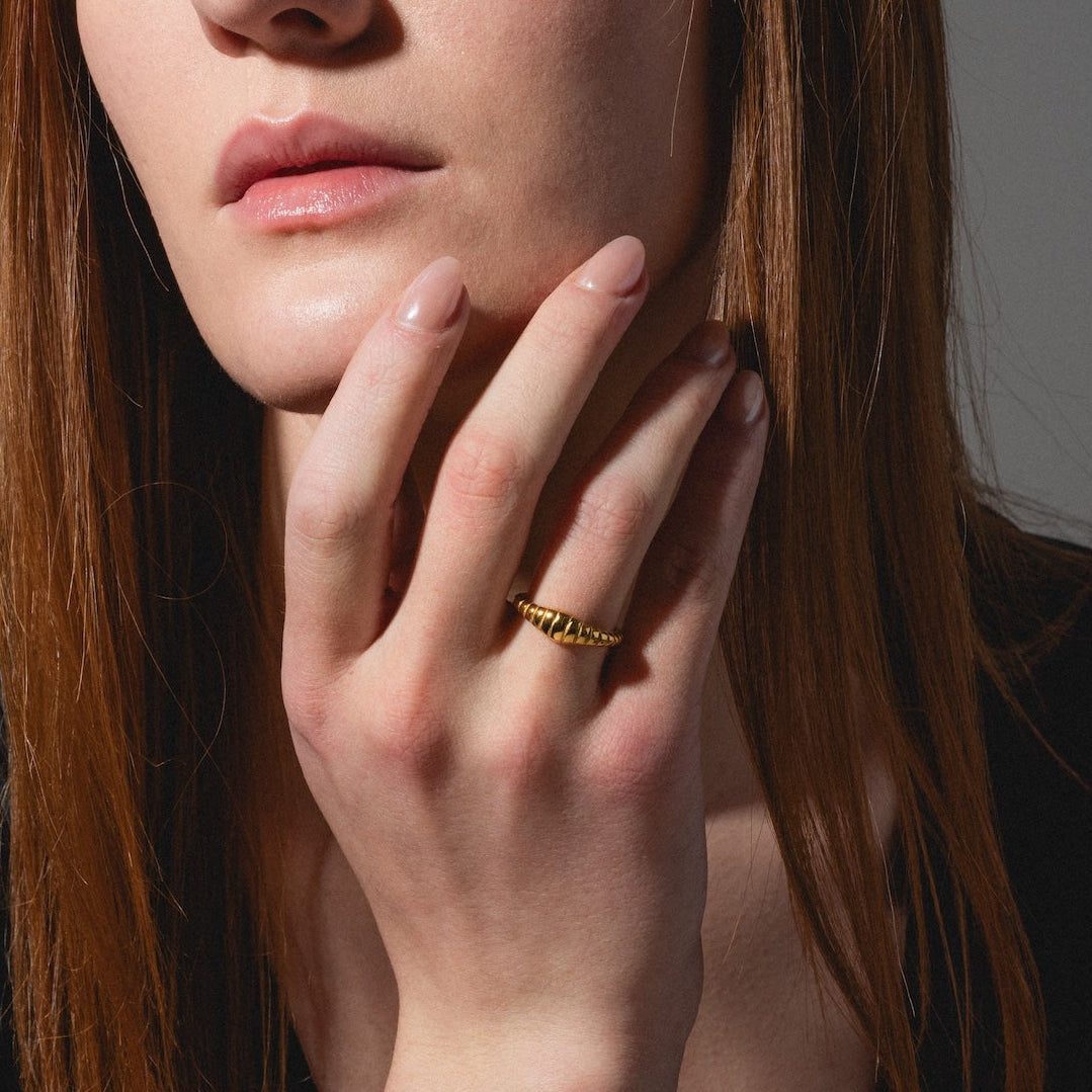 A young woman with long reddish-brown hair gazes to the side, resting her hand on her chin. She wears the Crew Ring and a black top, while soft natural light highlights her face.