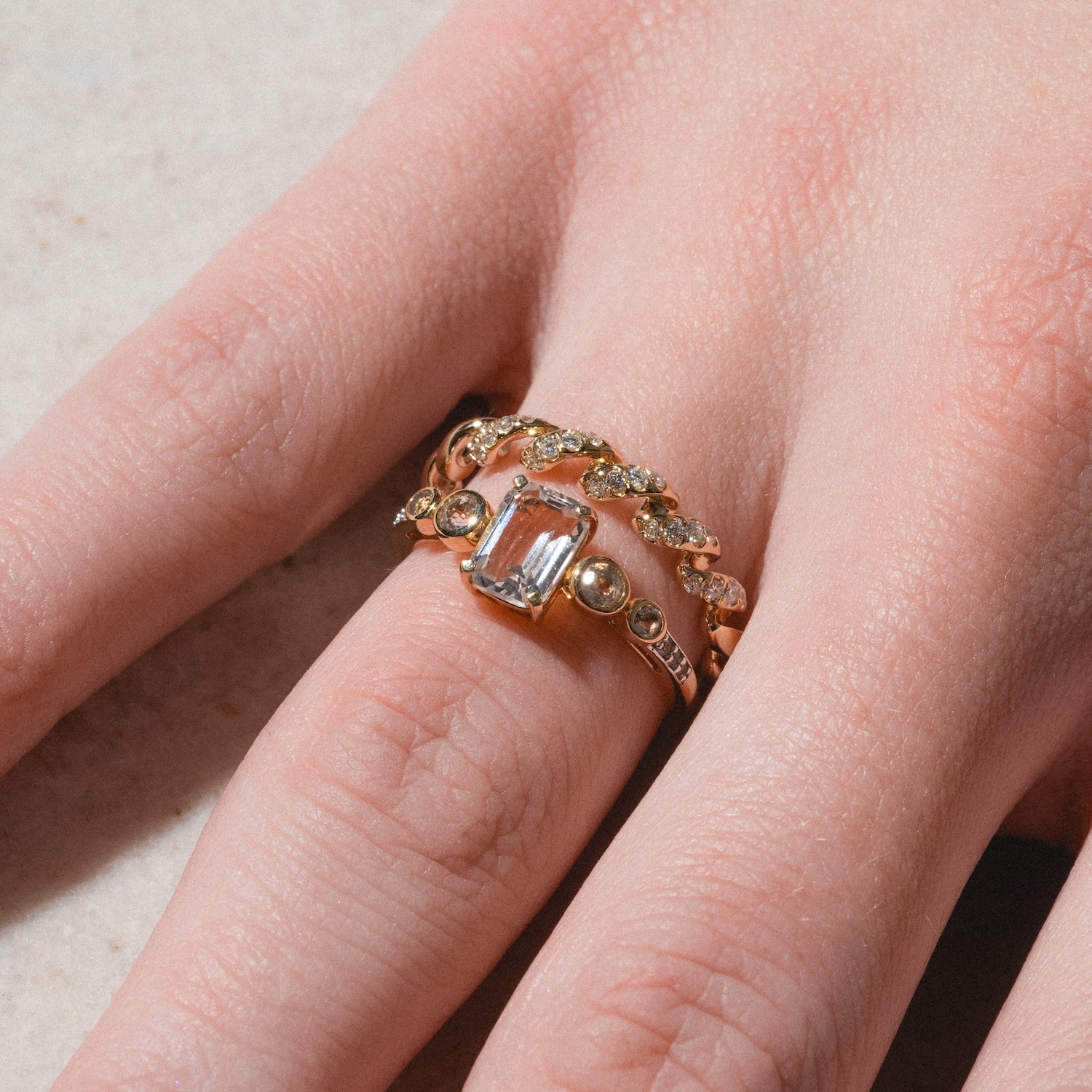 A close-up of a hand wearing two gold rings: one is a 14k solid yellow gold ring with a large rectangular clear gemstone, and the other is the 14k Fated Twist Diamond Ring featuring small diamonds along its twisted band.