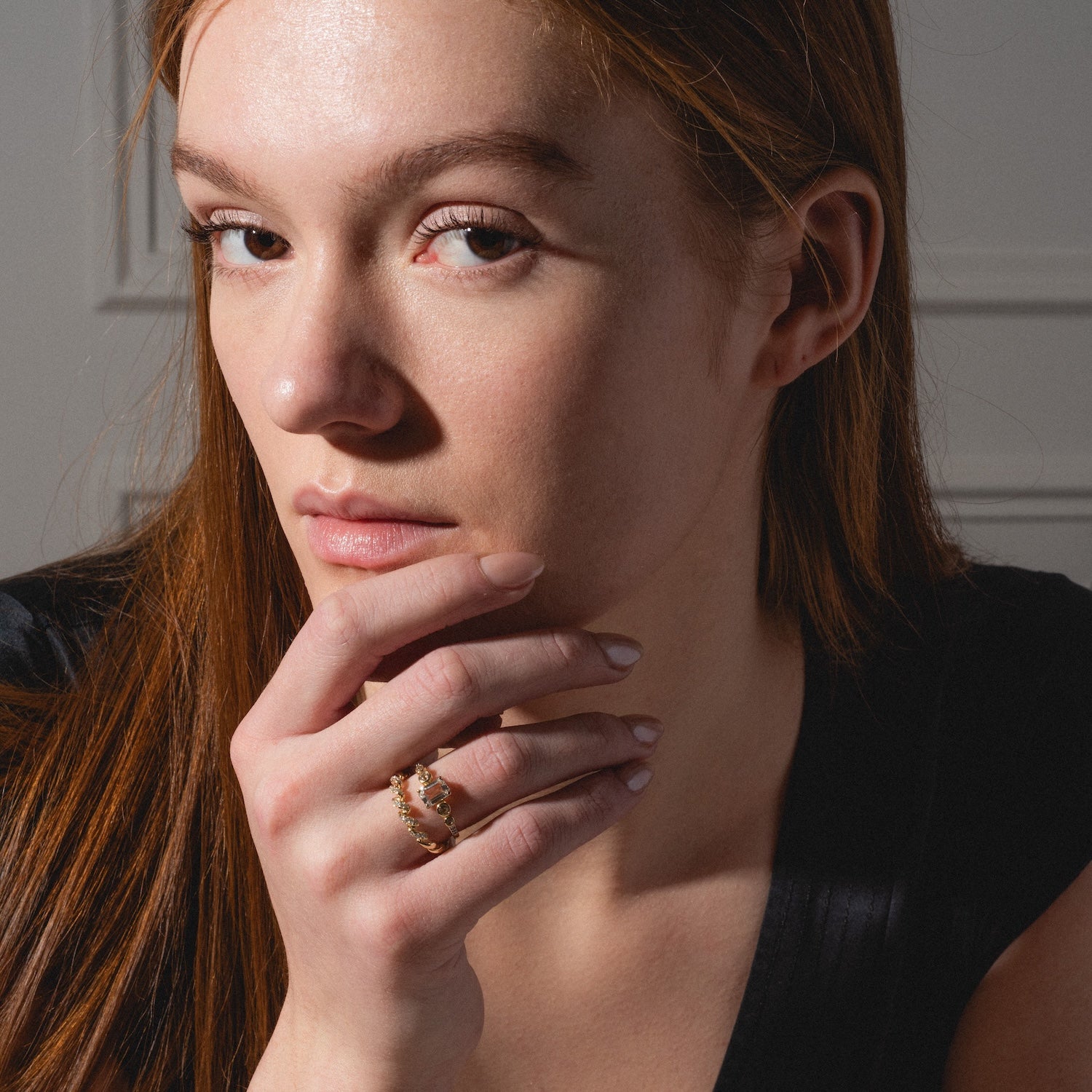 A woman with long, straight red hair and fair skin gazes thoughtfully at the camera, her hand on her chin. She wears the 14k Fated Twist Diamond Ring and a black top, soft light illuminating her face.