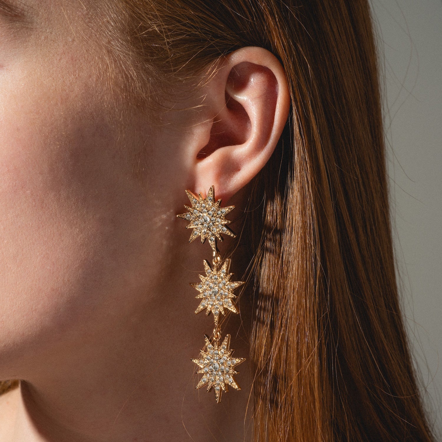 Close-up of a woman's ear wearing Celestial Drop Earrings, featuring three gold starburst shapes encrusted with sparkling clear stones, set against straight auburn hair.