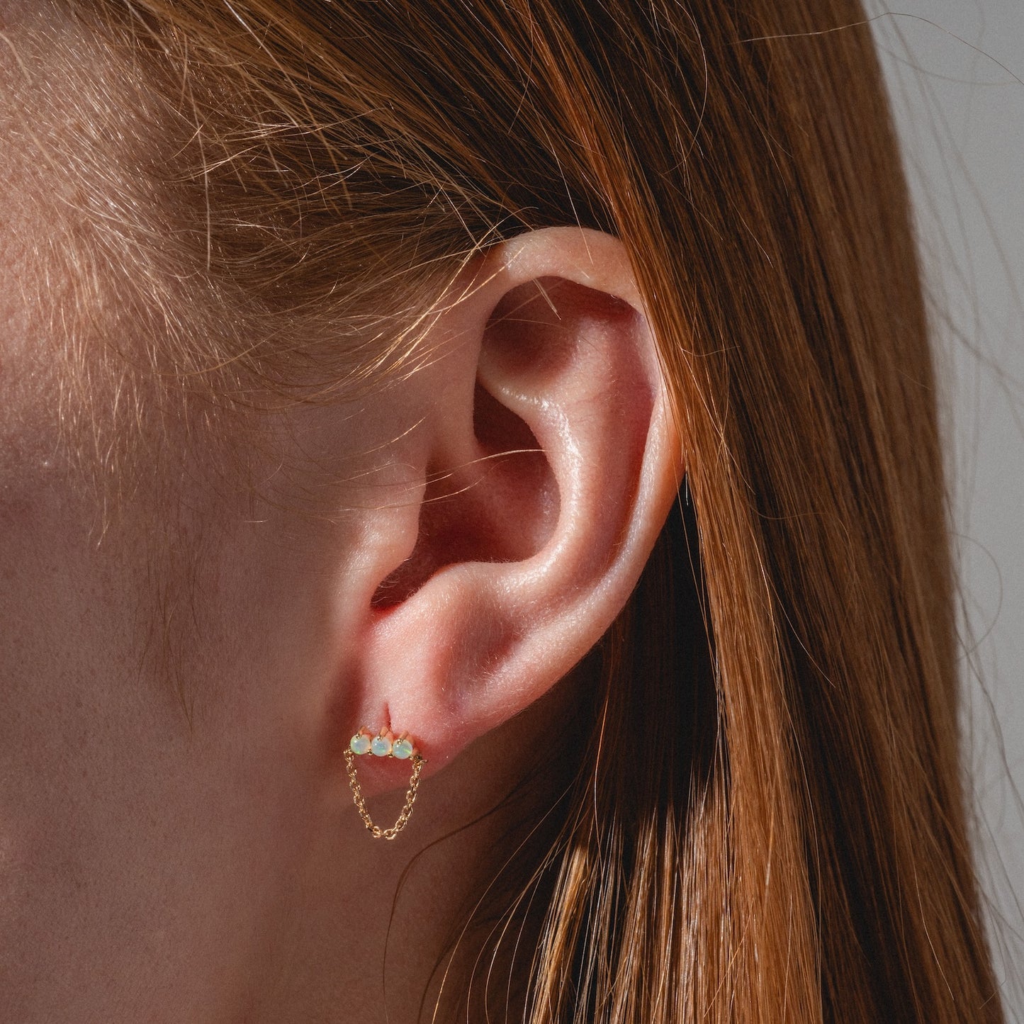 Close-up of a person with straight red hair wearing Solid Gold Opal Trio Chain Dangle Studs featuring three light blue stones and a delicate gold chain—ideal for those who love unique opal earrings.