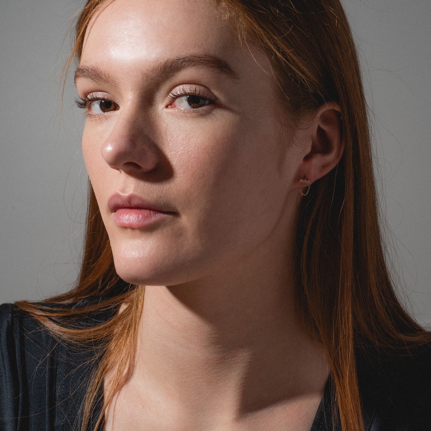 A woman with long, straight reddish-brown hair and fair skin gazes confidently at the camera, wearing a black top and elegant Solid Gold Opal Trio Chain Dangle Studs. Soft lighting highlights her understated style.