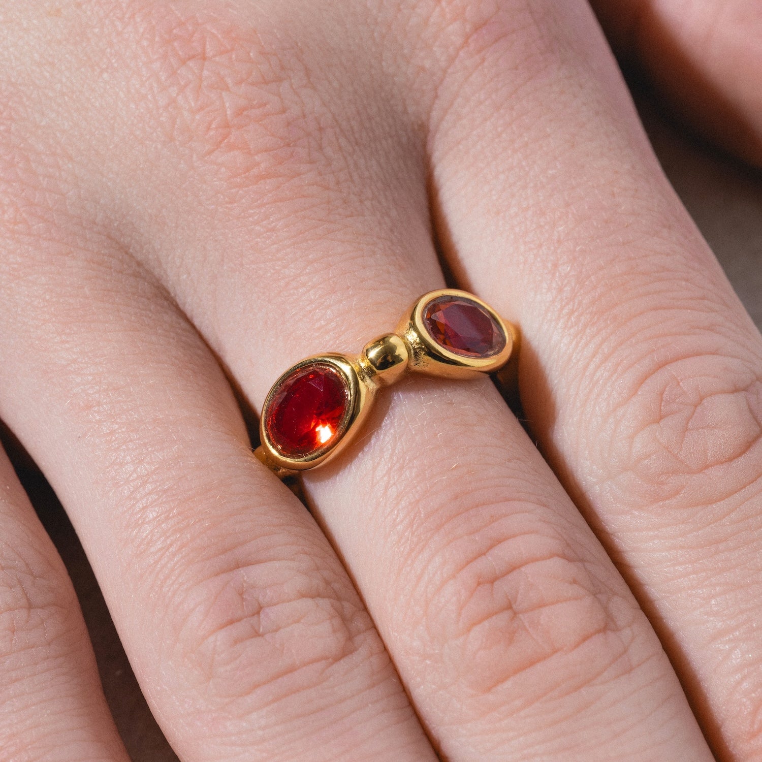 The Bita Ring is a gold ring worn on a person's finger, featuring two oval-shaped red gemstones set side by side and connected by a gold bead in a unique design.