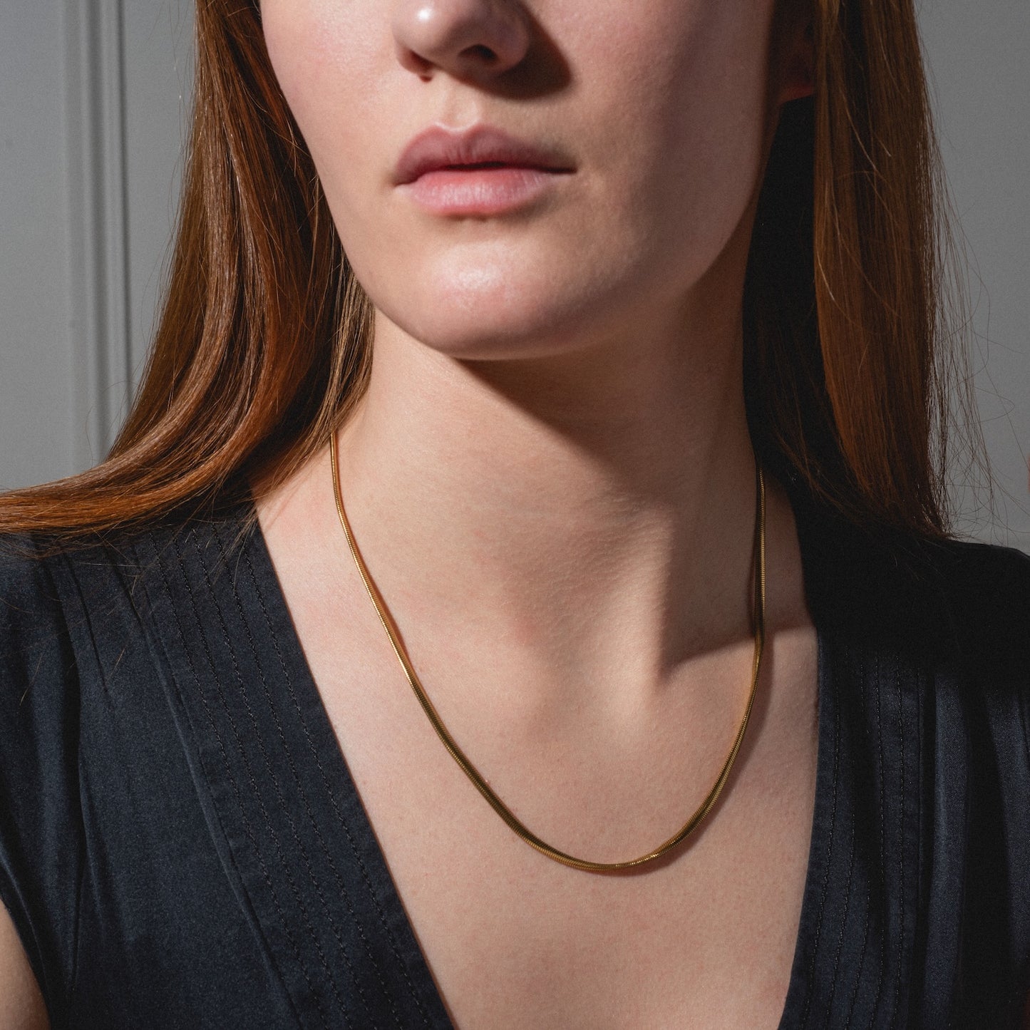 A woman with long brown hair wears the Classic Snake Chain Necklace, styled with a black V-neck top, shown from the shoulders up against a neutral background.