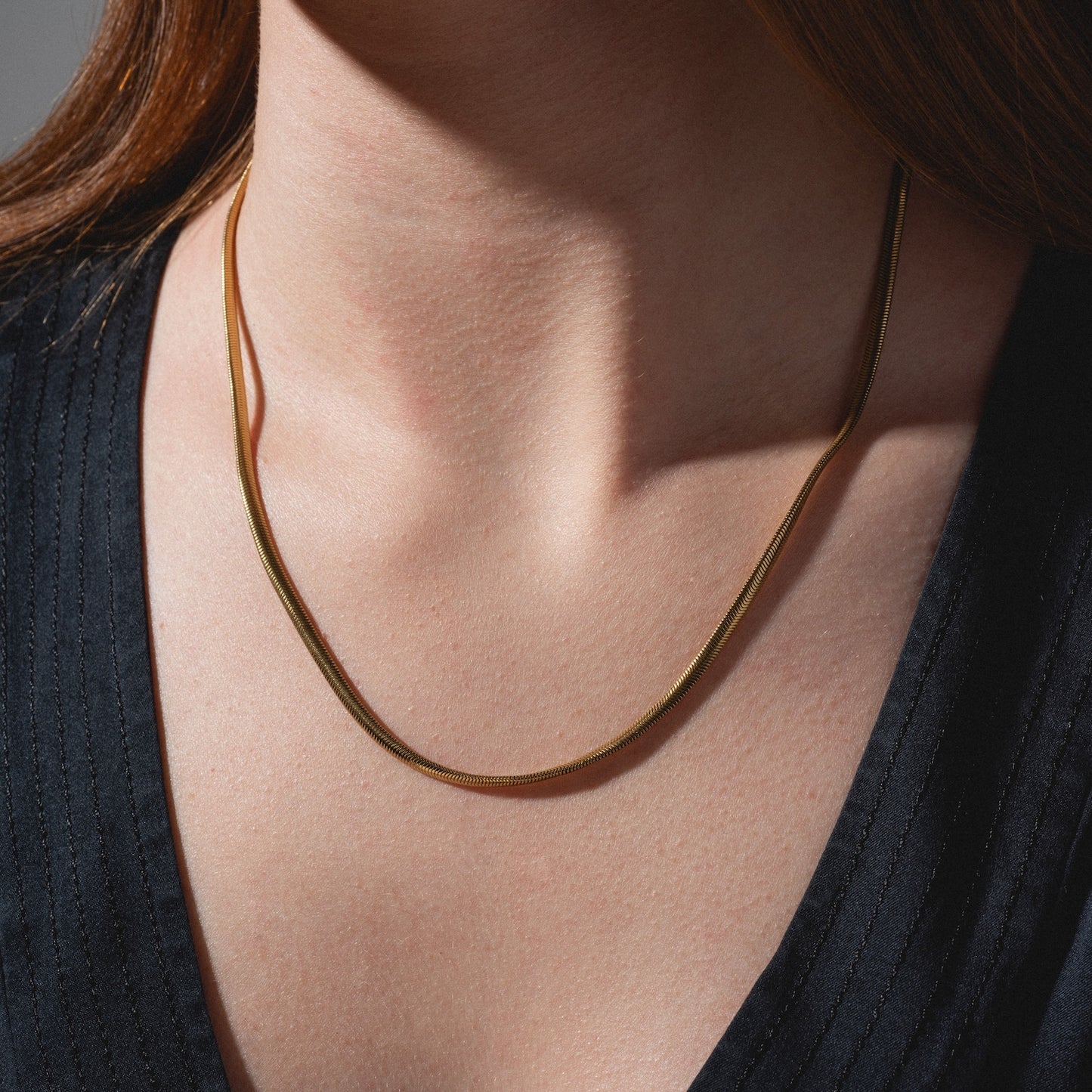 A close-up of a person wearing the Classic Snake Chain Necklace, a thin gold-plated piece styled over a dark V-neck top, with brown hair partially visible on the left side.