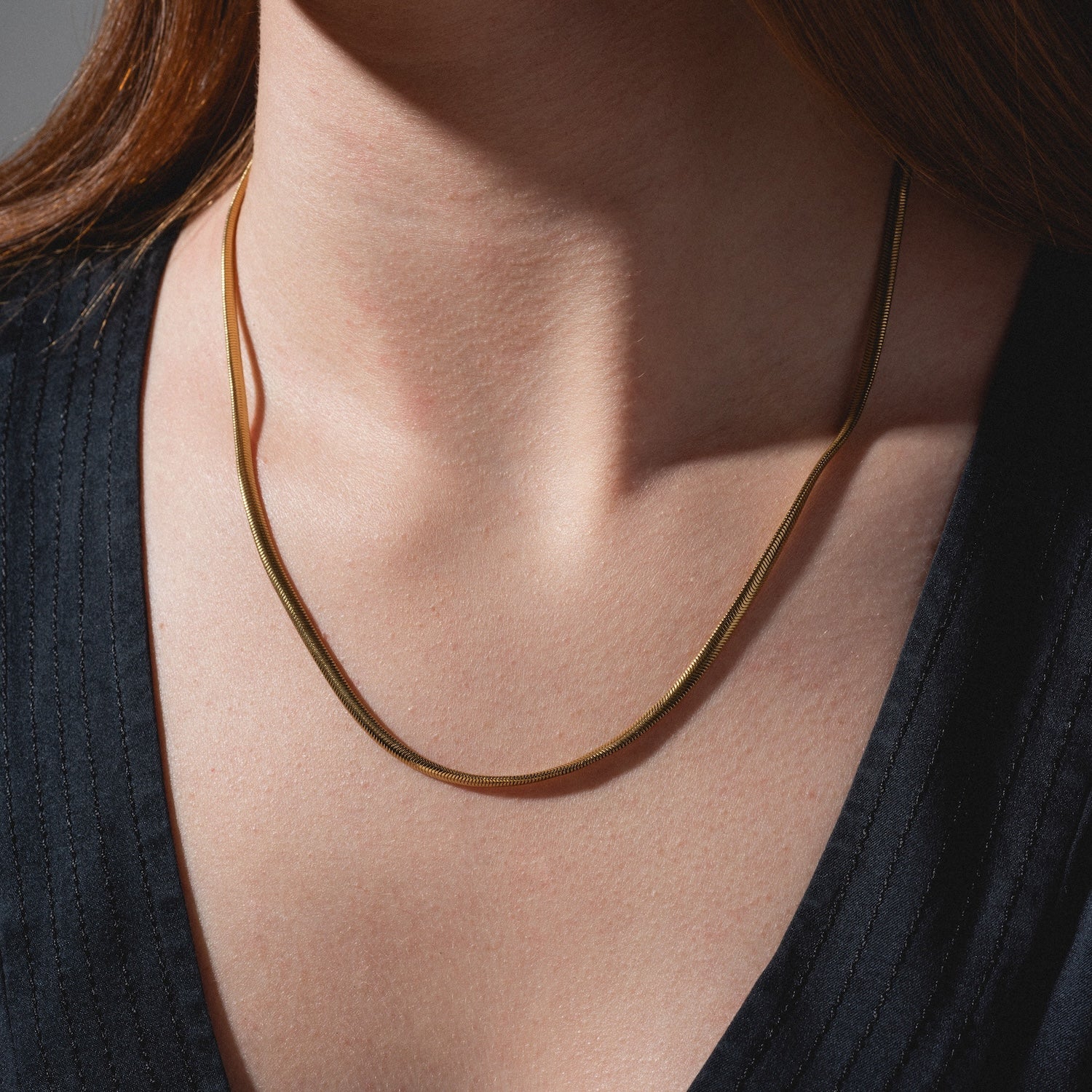 A close-up of a person wearing the Classic Snake Chain Necklace, a thin gold-plated piece styled over a dark V-neck top, with brown hair partially visible on the left side.