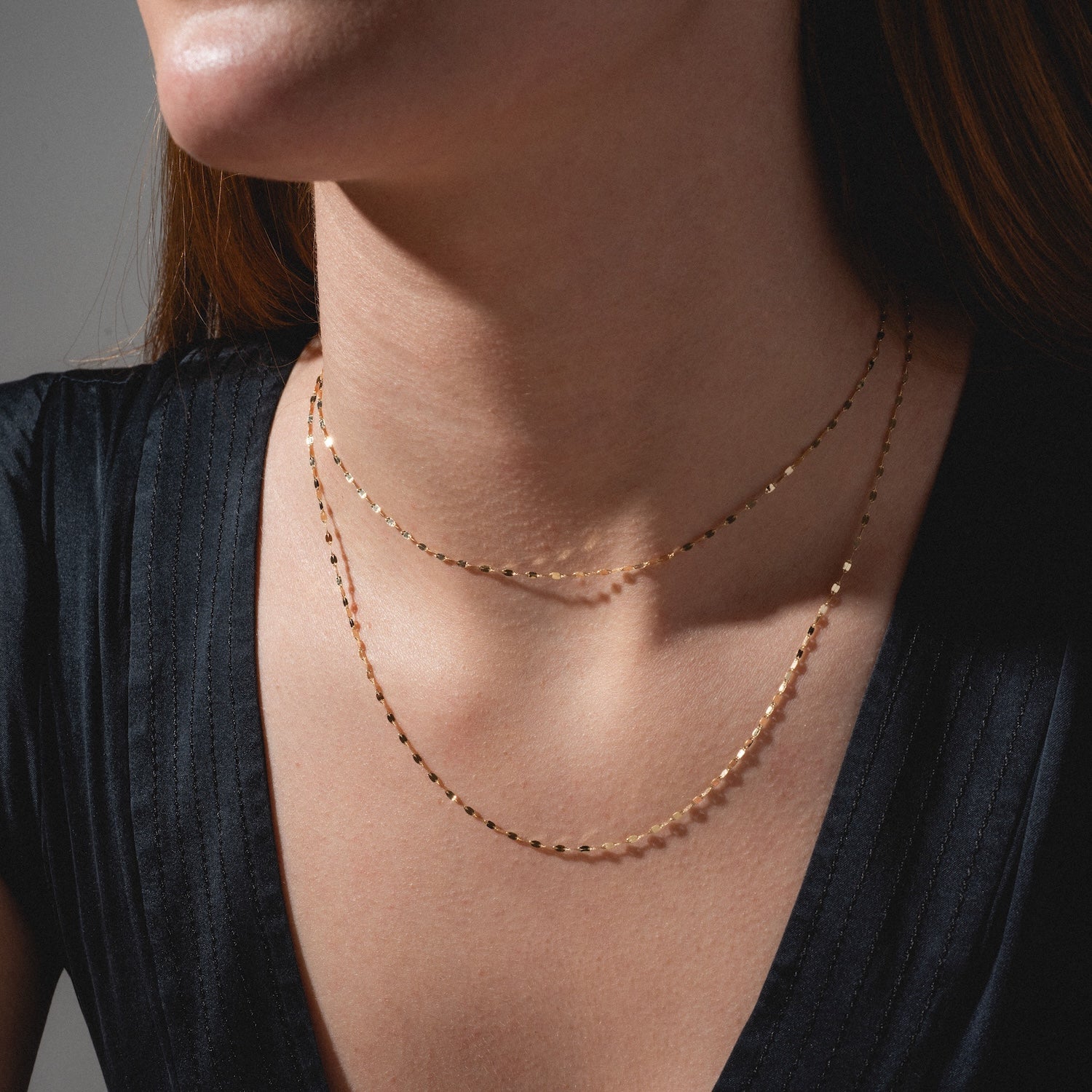 A woman showcases her neckline in a black V-neck top, wearing the Solid Gold Chain for Charms layered with a delicate charm necklace. The elegant gold jewelry highlights her collarbone.