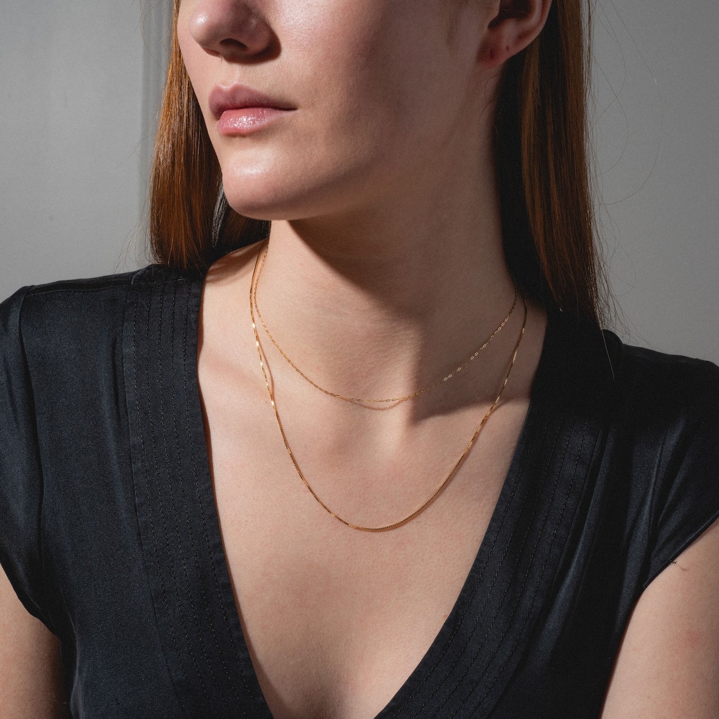 A woman with straight reddish-brown hair wears a black V-neck top and two gold necklaces—one is the Solid Gold Box Chain in 9k solid gold. Her face is partly visible, set against a plain, softly lit background.