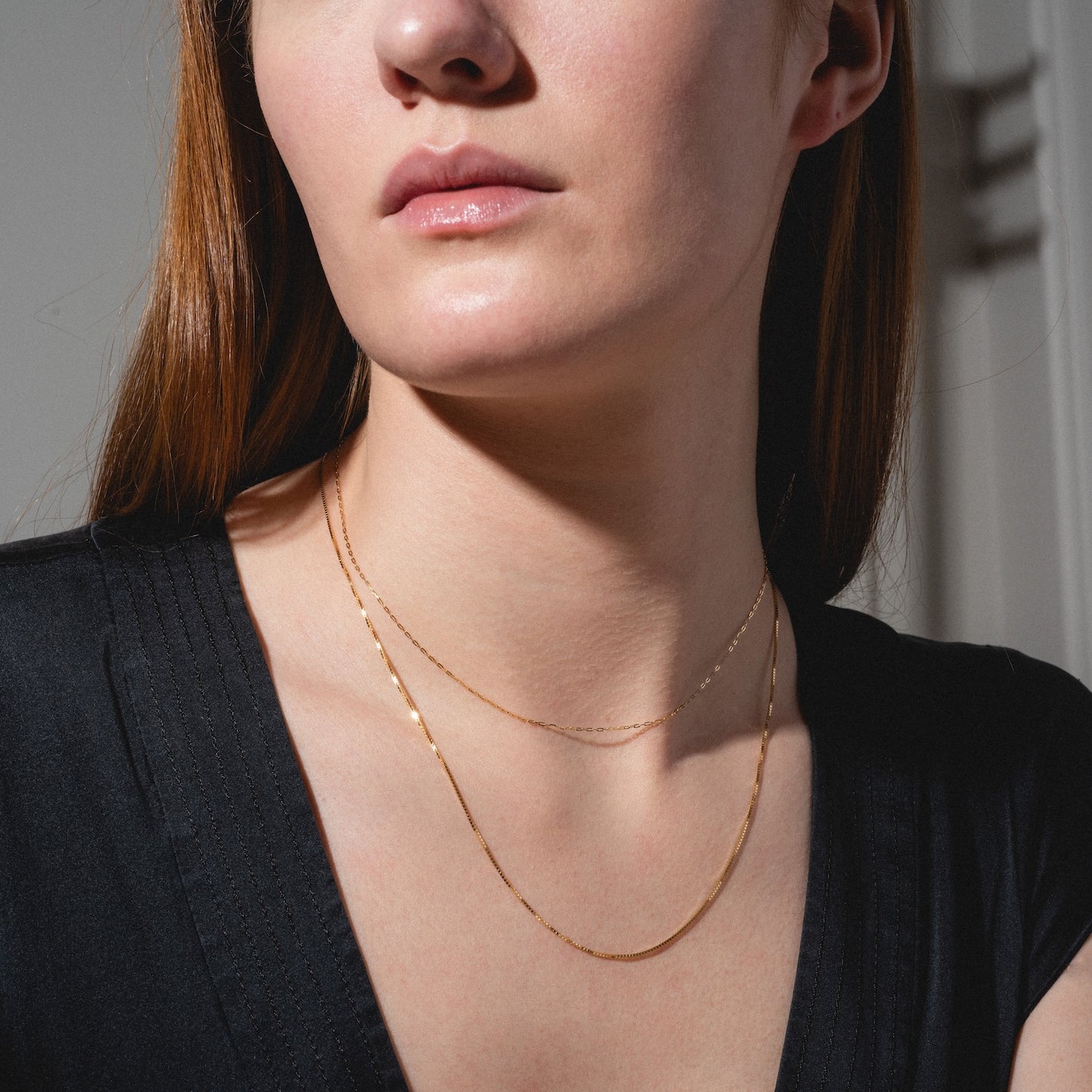A woman with long auburn hair wears a black V-neck top and two gold necklaces, one of which is the Solid Gold Delicate Paperclip Chain. The image is cropped to show her face from the nose down, neck, and upper chest.