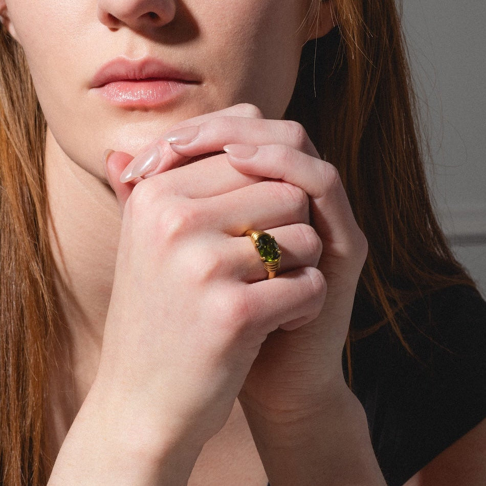 A woman with long auburn hair, wearing a black top and the Stone Statement Ring featuring a green gemstone, rests her chin on her hands and gazes thoughtfully to the side against a softly lit neutral background.