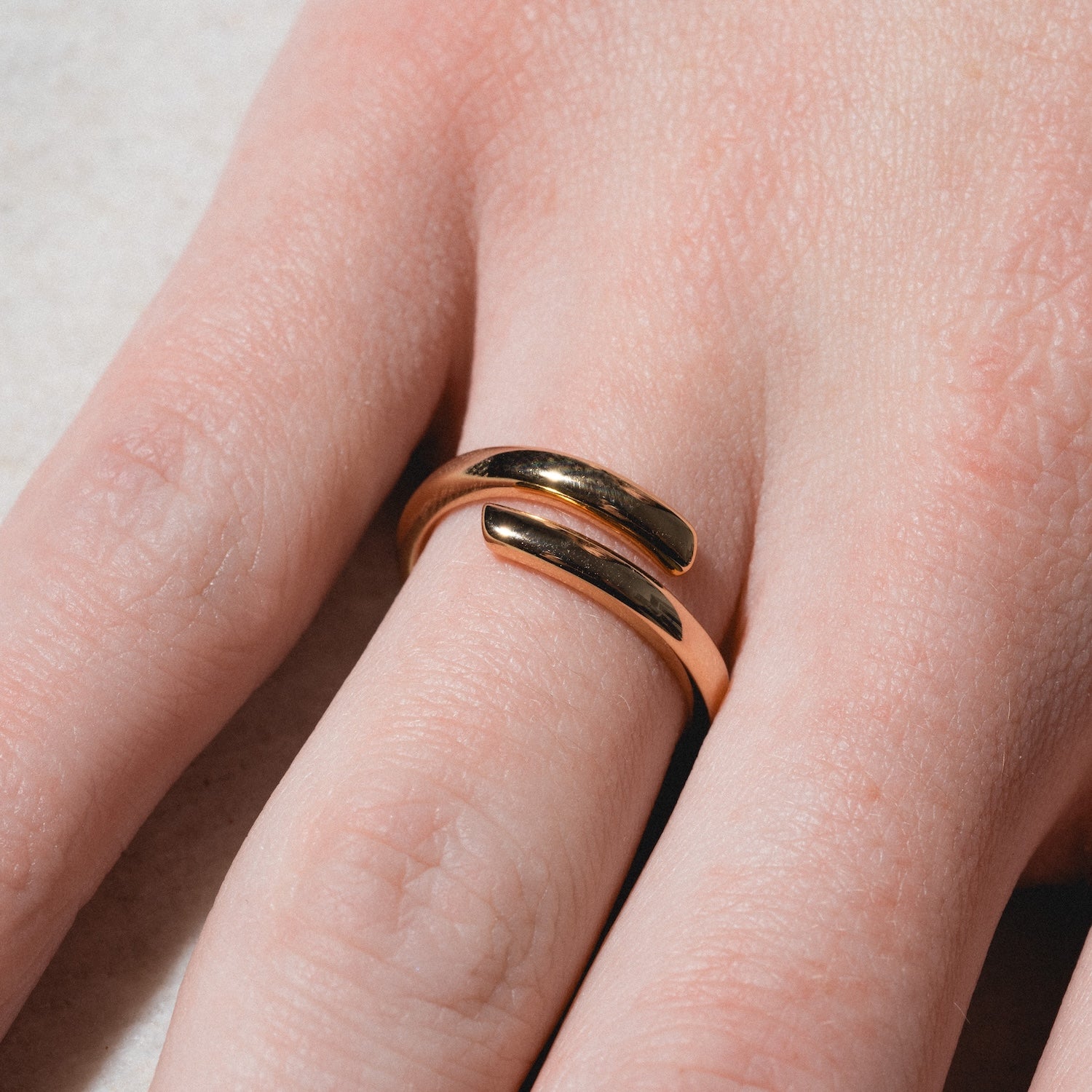 A hand wears the Wrap Around Ring, a gold minimalist open ring with two curved ends facing each other on the middle finger, against a light, neutral background.