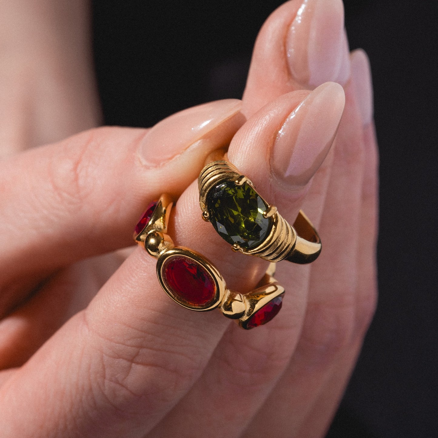 A manicured hand holds the Stone Statement Ring—one gold band with a large green gemstone, and another with two red oval gems—against a dark background.