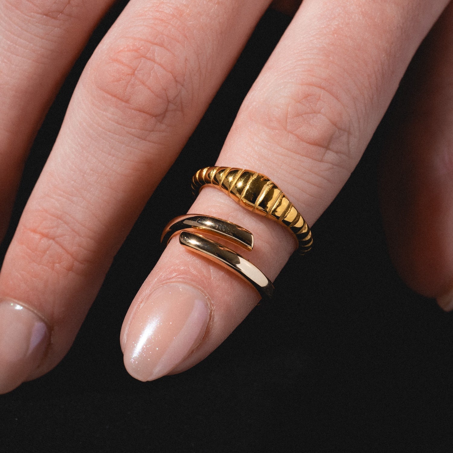 A close-up of a hand with neatly manicured nails wearing two gold rings, including the Crew Ring with its ridged design and another smooth, curved ring, against a black background.