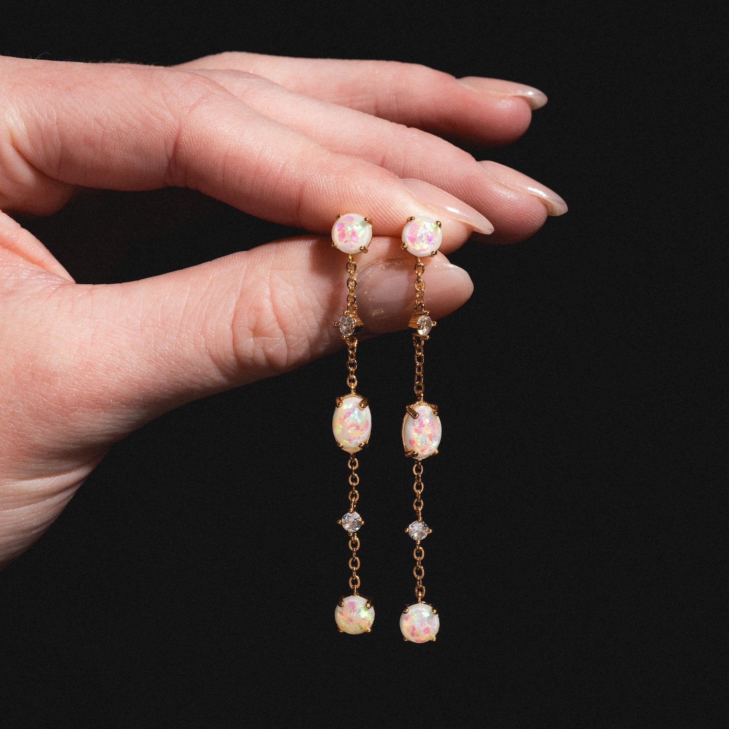 A hand holds the Elegant Opal Sway Dangle Earrings—delicate gold drops with pink and white floral beads and diamond-shaped crystals—shown against a black background.