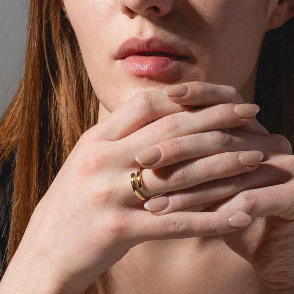 A woman with long reddish-brown hair and natural makeup gazes forward with her hands clasped under her chin, wearing the Wrap Around Ring and a dark top against a neutral background.