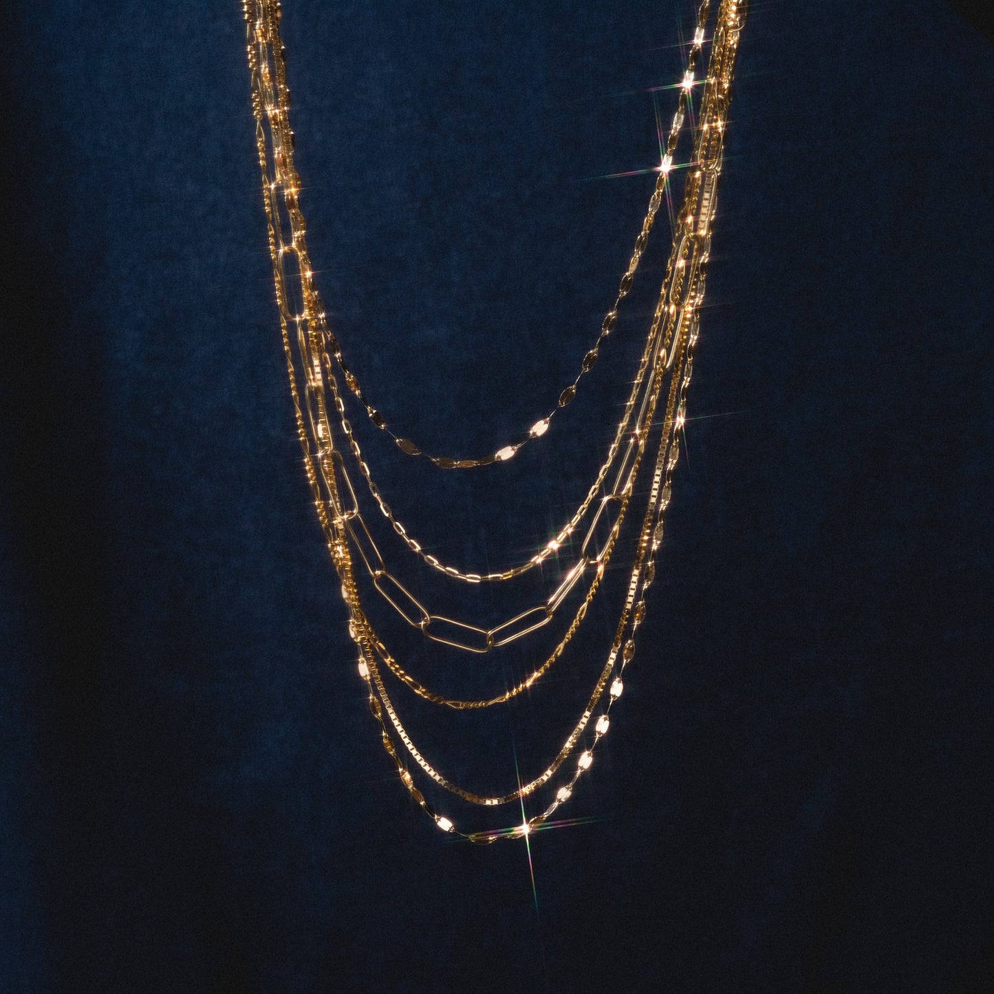 The Solid Gold Chain for Charms shines against a deep blue backdrop, its layered styles reflecting light and sparkling—making it the ideal foundation for your personalized charm necklace.