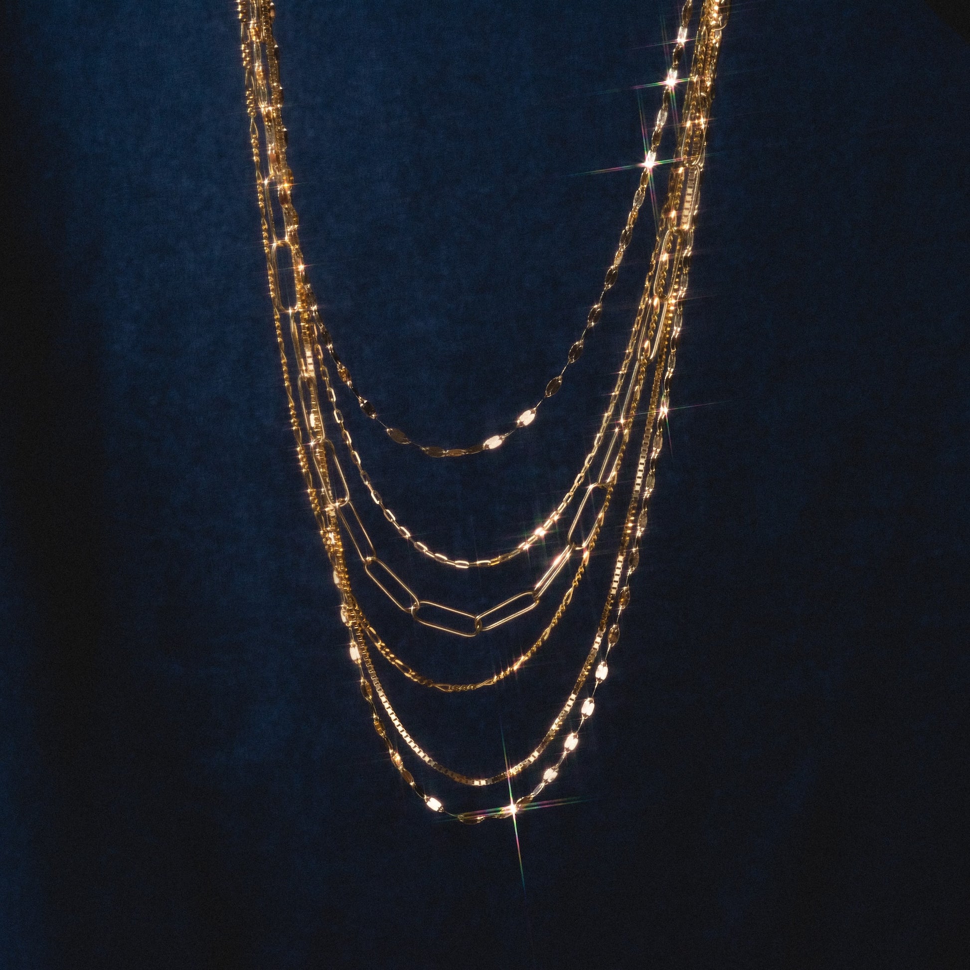 The Solid Gold Chain for Charms shines against a deep blue backdrop, its layered styles reflecting light and sparkling—making it the ideal foundation for your personalized charm necklace.