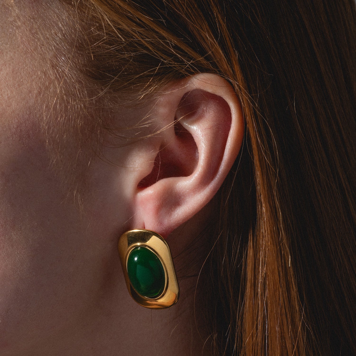 A close-up of a person’s ear wearing Ashley Earrings: large gold earrings with an oval green stone. Straight auburn hair is partially visible.