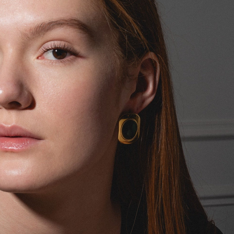 A woman with long brown hair, wearing a black top and the Ashley Earrings—gold squares—gazes calmly at the camera, her features highlighted by soft lighting against a neutral background.