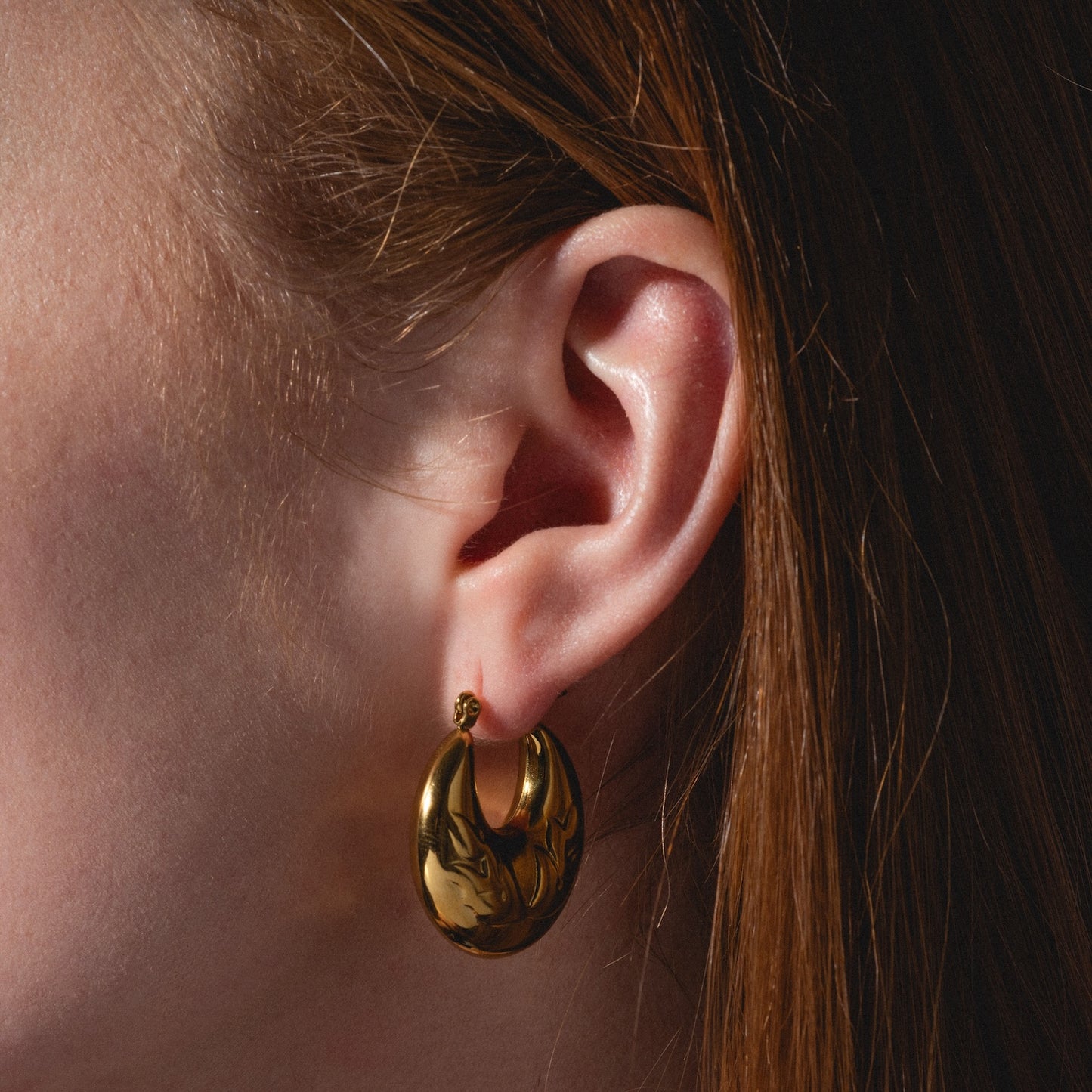 Close-up of a person with long auburn hair wearing shiny, gold Goldie Earrings—oval-shaped hoops featuring a subtly marbled, wavy design.