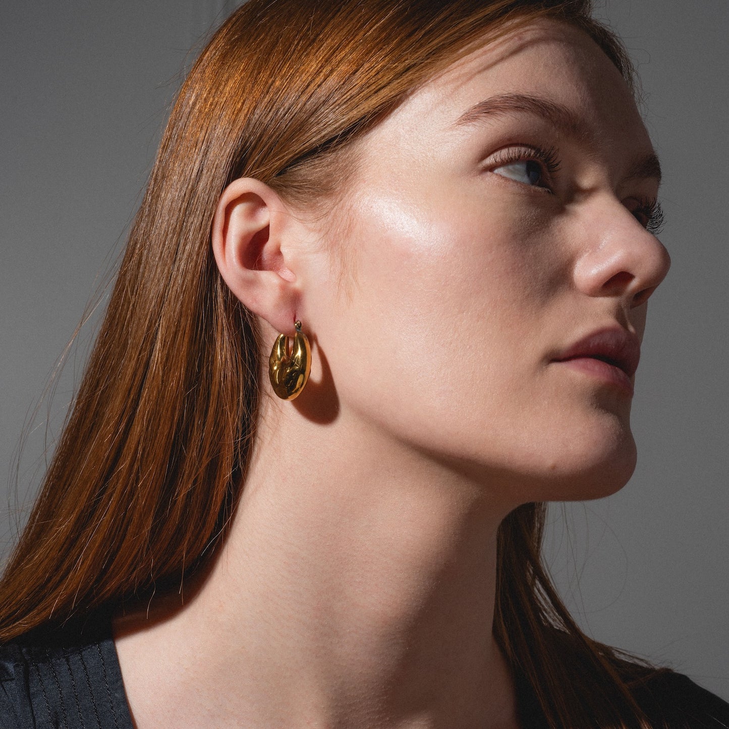 A woman with long, straight, reddish-brown hair wears a black top and shiny Goldie Earrings, gazing slightly upwards and to the side in close-up lighting that enhances her clear skin.