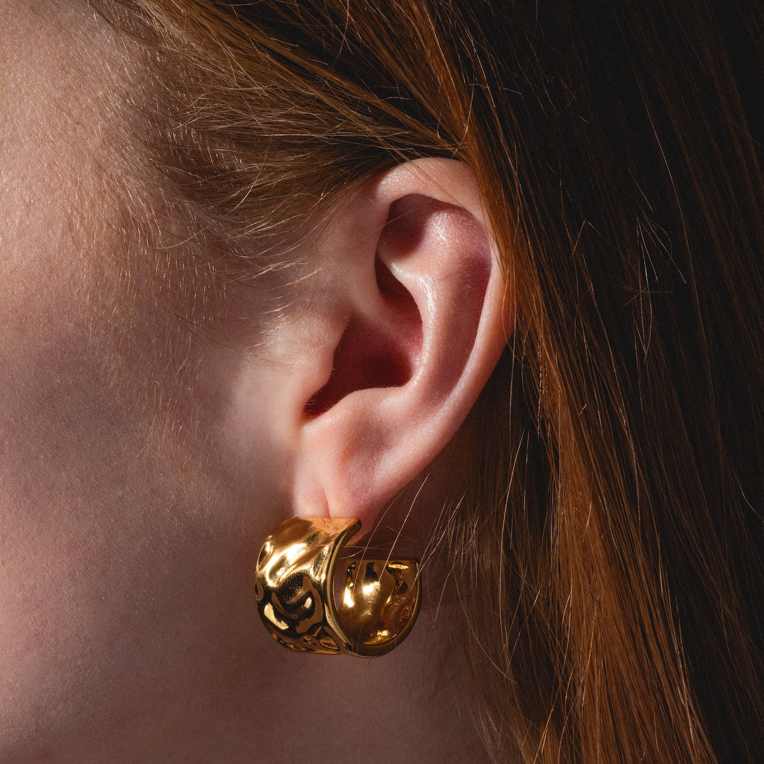 Close-up of a person with light skin and auburn hair wearing the Lasso Hoops—large, shiny gold earrings featuring an intricate, sculpted design. The focus is on the ear and the earring.