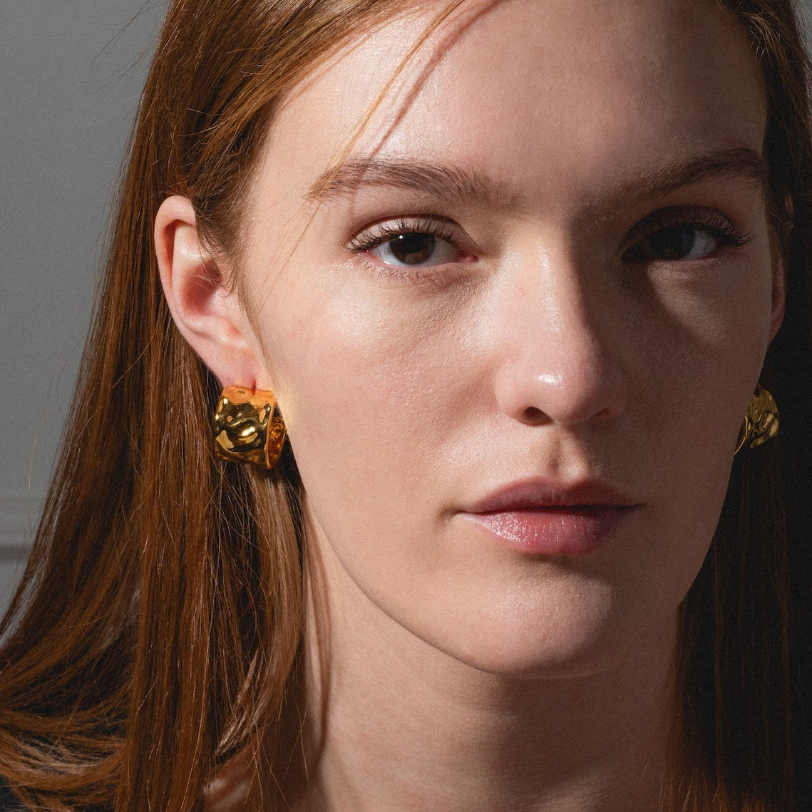A woman with straight, long auburn hair stares confidently at the camera, wearing Lasso Hoops and a black top, her face softly highlighted by natural light.