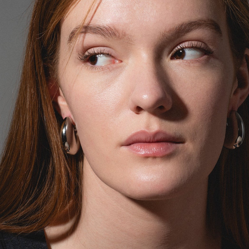 A woman with long, straight auburn hair looks to her left, wearing Remy Earrings—large silver hoops—with a black top. The lighting highlights her clear skin and minimal makeup against a light-colored wall.