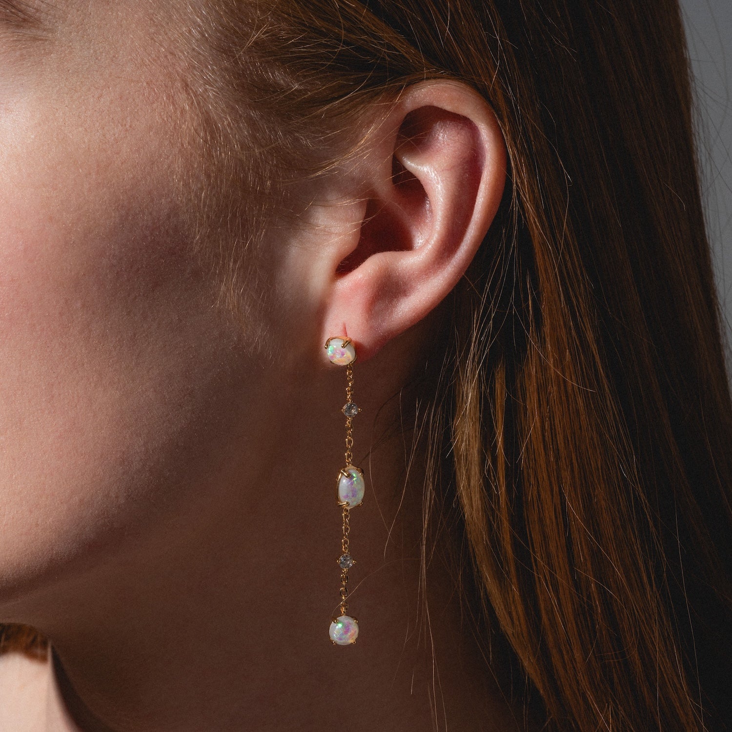 Close-up of a woman's ear wearing Elegant Opal Sway Dangle Earrings with three iridescent opal gemstones. Her brown hair is tucked behind her ear, and the neutral background highlights the gold and opal details.