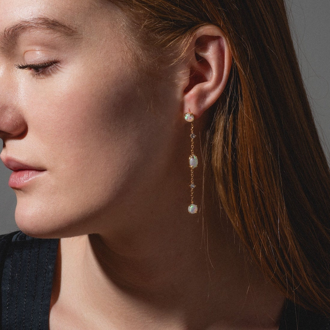 A woman with long auburn hair and closed eyes wears a black top and Elegant Opal Sway Dangle Earrings. The long gold earrings with white pearl accents are showcased in profile against a neutral background.