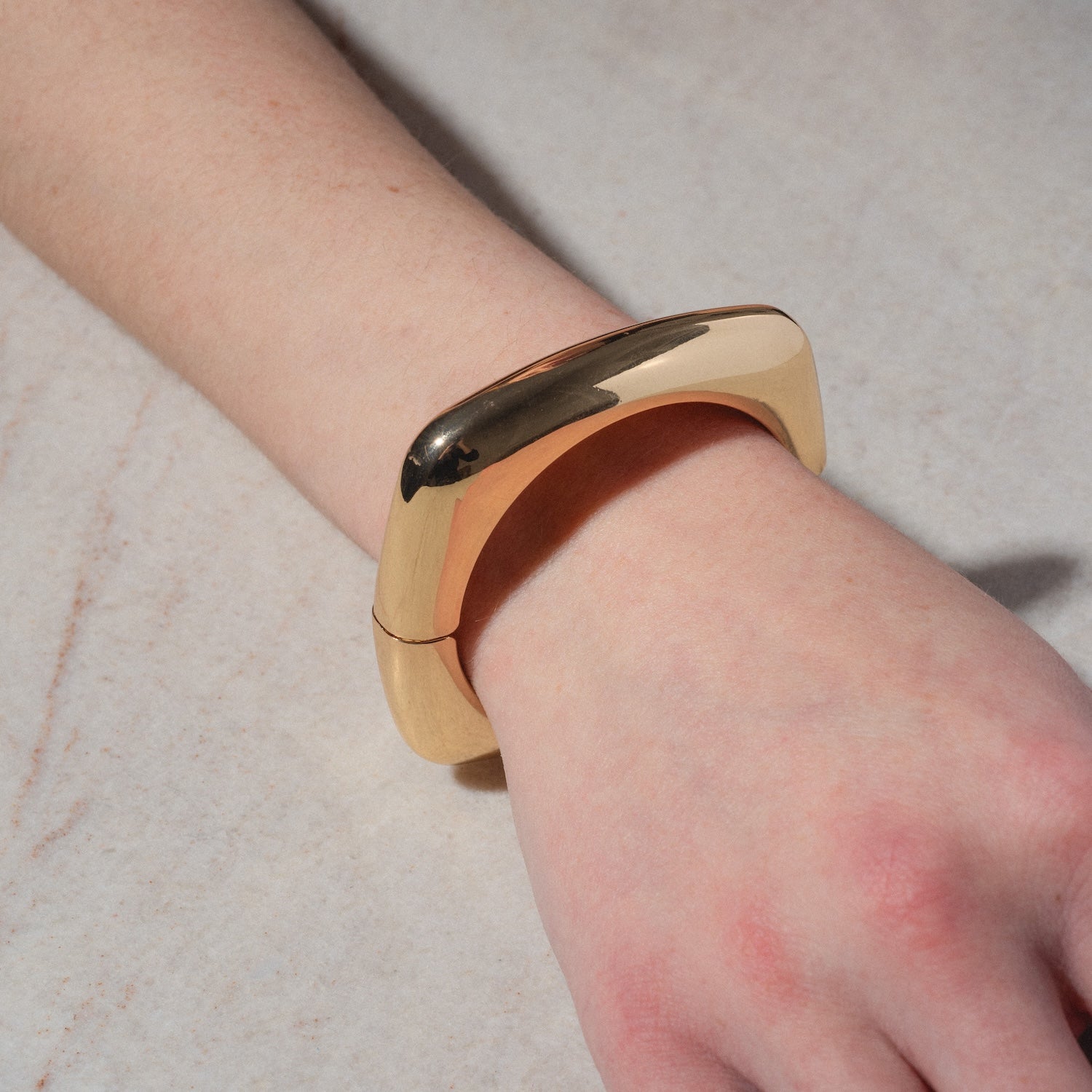A hand rests on a light, textured surface, wearing the Square Cuff Bracelet—a chunky gold piece with curved edges and a smooth, polished finish.